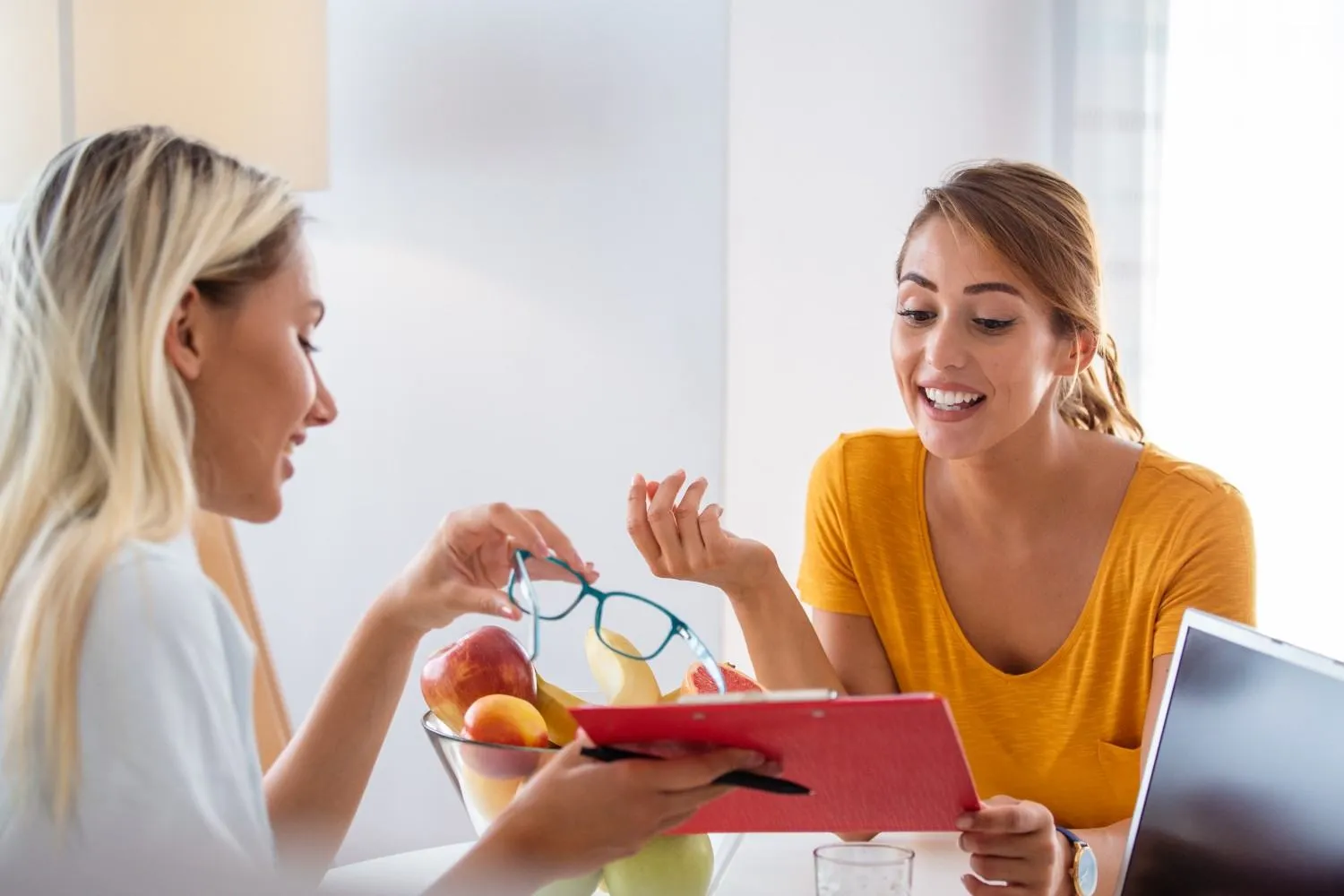 Two women talking over a bowl of fresh fruit while reading notes on a clipboard